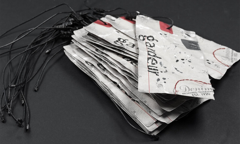 Stapel mehrerer Karton-Hangtags im Vintage Used-Look mit rotem Akzentdruck, partieller Silberfolienprägung und schwarzem Plombenfaden für eine Denim-Marke – individuelle Hangtag-Produktion von WEROCA Kartonagen Bielefeld