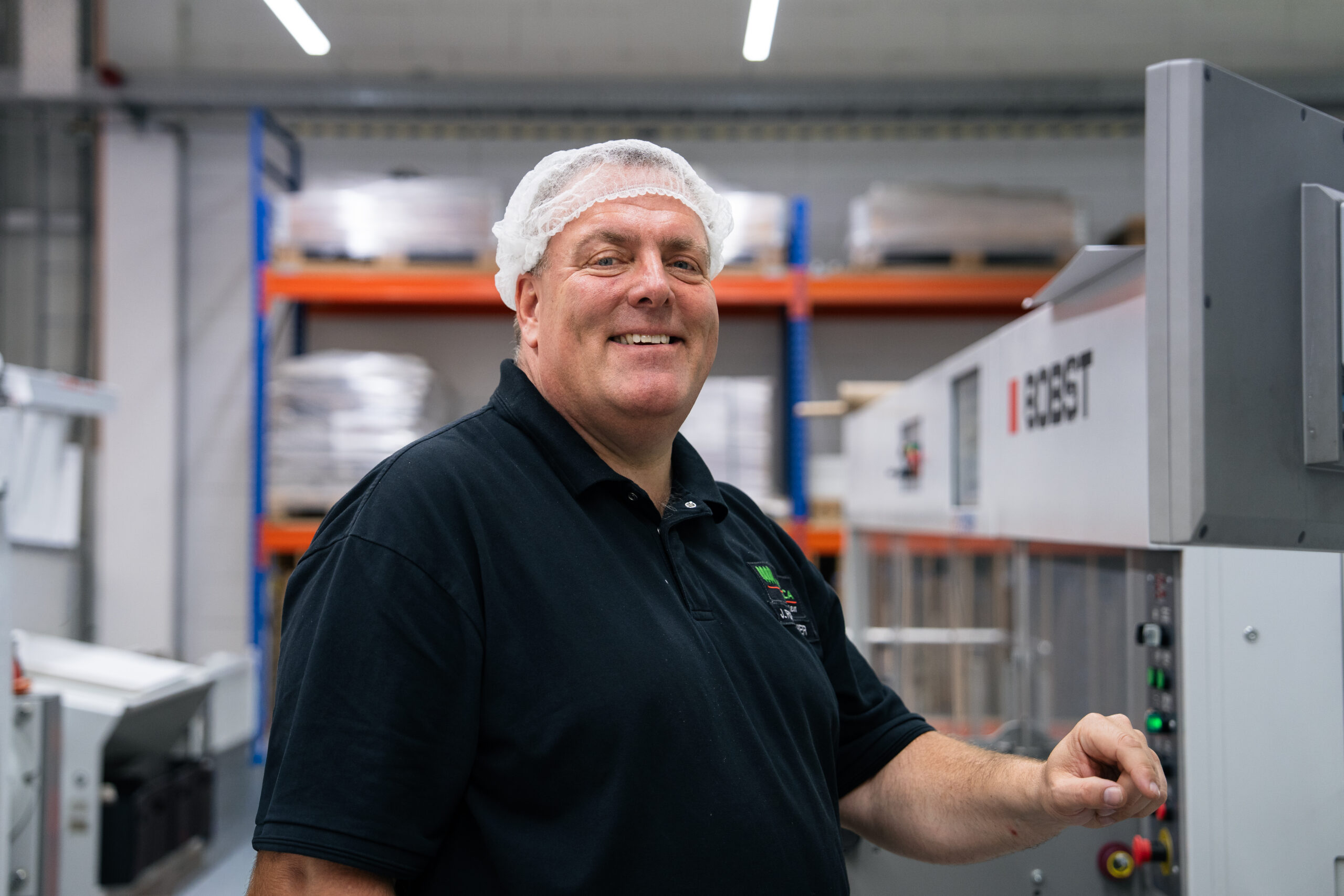 Lächelnder WEROCA-Mitarbeiter mit Haarnetz an einer Bobst Stanzmaschine in der Produktionshalle in Bielefeld-Hillegossen – Fachkompetenz und Qualität in der Kartonverpackungsherstellung