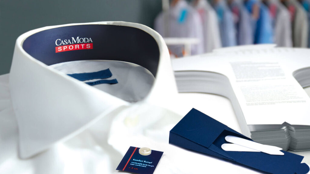 Weißes Business-Hemd mit dunkelblauem Nackenbändchen, Krageneinlagen, Kragensteg und bedrucktem Pocketflasher – maßgefertigte Trimmings für die Hemdenbranche von WEROCA Kartonagen Bielefeld