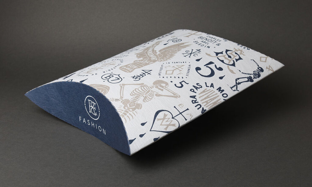 Weiße Kissenverpackung mit vollflächigem Allover-Druck in Blau und Beige und dunkelblauem Verschluss mit Markenlogo für eine Fashion-Marke – individuelle Textilverpackungen von WEROCA Kartonagen Bielefeld