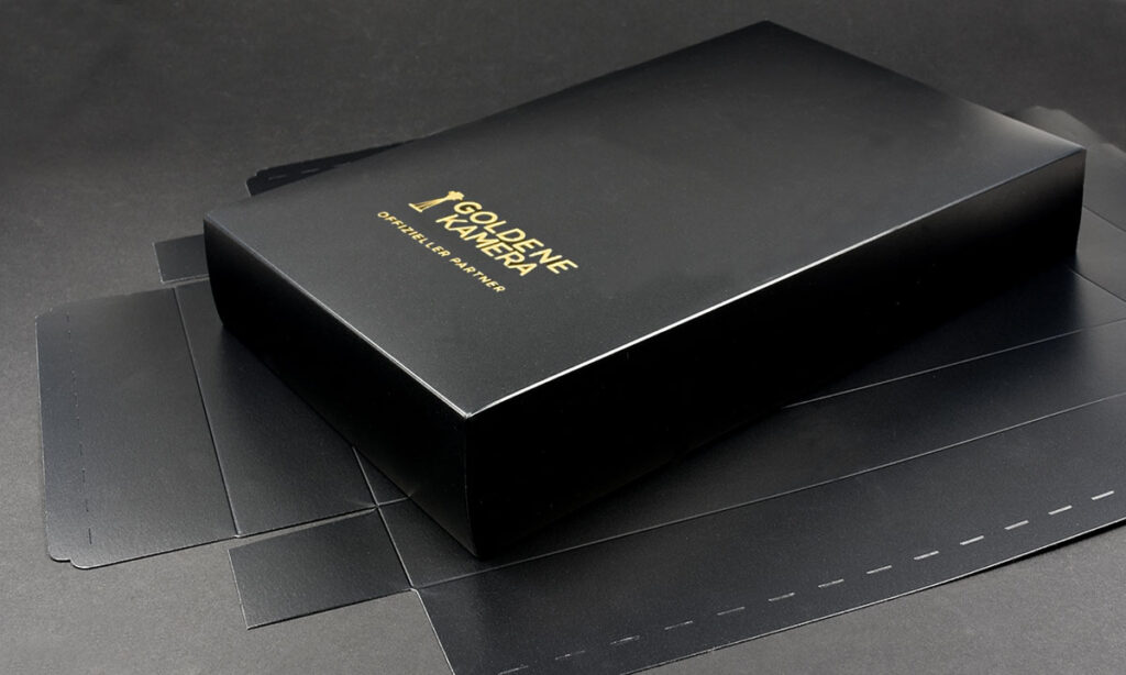 Schwarze Faltschachtel mit Goldfolienprägung „Goldene Kamera – Offizieller Partner" auf flachem Stanznutzen liegend – hochwertige Event-Verpackung mit Heißfolienveredelung von WEROCA Kartonagen Bielefeld