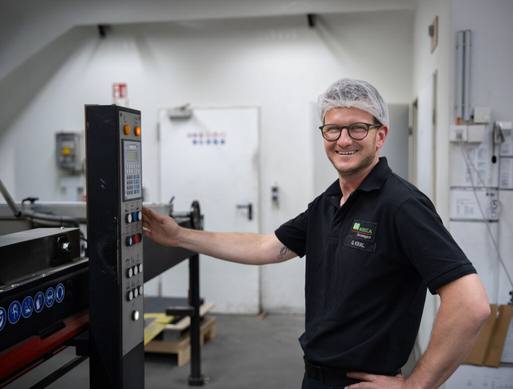 Lächelnder WEROCA-Mitarbeiter bedient das Steuerungspanel einer Stanzmaschine in der Produktionshalle der WEROCA Kartonagen GmbH & Co. KG in Bielefeld-Hillegossen