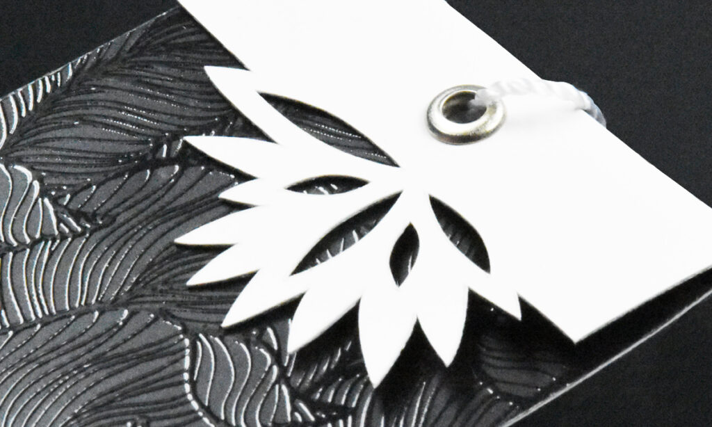 Weißes Hangtag von WEROCA Kartonagen Bielefeld mit ausgestanztem Blattmotiv über gemustertem Untergrund, Detailaufnahme für Fashion & Textil-Hangtags.