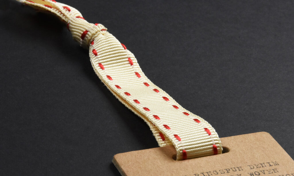 Cremefarbenes Ripsband mit roten Nähten von WEROCA Kartonagen Bielefeld an einem braunen "Ringspun Denim"-Hangtag, Detailaufnahme für Fashion & Textil-Hangtags.
