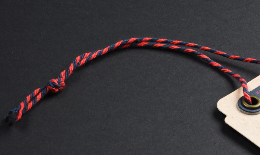 Rot-blau gedrehte Kordel von WEROCA Kartonagen Bielefeld mit Knoten an einem beigen Hangtag mit Messingöse, Detailaufnahme für Fashion & Textil-Hangtags.