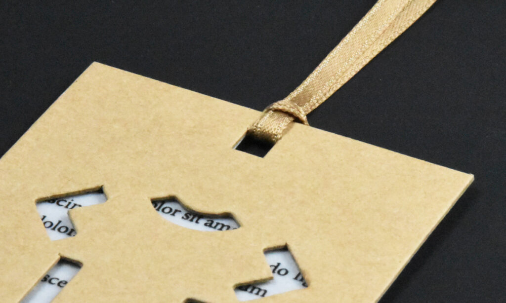 Hangtag von WEROCA Kartonagen Bielefeld mit ausgestanzten Buchstaben über Textpapier und goldenem Satinband, Detailaufnahme für Fashion & Textil-Hangtags.