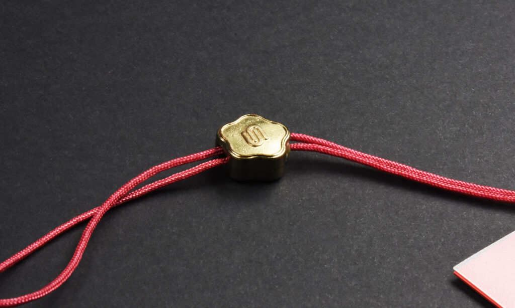 Blütenförmiger Goldclip mit "S"-Prägung von WEROCA Kartonagen Bielefeld an roter Kordel, Detailaufnahme für Fashion & Textil-Hangtags.