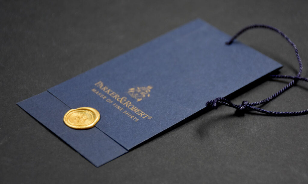 Dunkelblaues Karton-Hangtag mit goldenem Aufdruck, goldenem Wachssiegel und blauer Seidenkordel für eine Premium-Herrenhemden-Marke – exklusive Hangtag-Veredelung von WEROCA Kartonagen Bielefeld