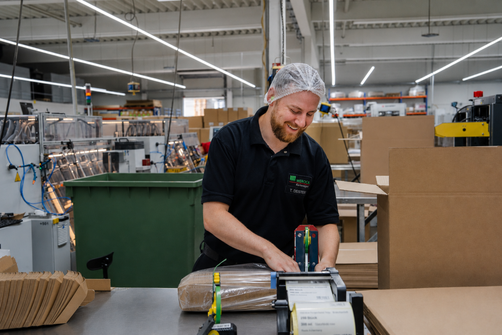 Lächelnder WEROCA-Mitarbeiter mit Haarnetz an einem Packtisch an einer Klebemaschine, der Kartonschalen in Polybeutel unterverpackt, in der Produktionshalle in Bielefeld-Hillegossen – Fachkompetenz und Qualität in der Kartonverpackungsherstellung